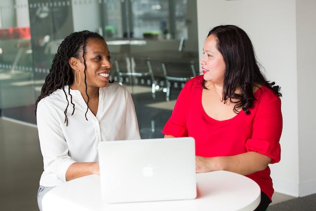 two people sitting at table talking with laptop in front of them