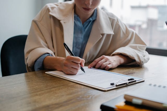 person sitting at a table writing on a piece of paper clipped on a clipboard